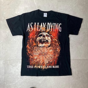 May include: Black t-shirt featuring the band name "AS I LAY DYING" in white, above a graphic of a skull and a fiery, abstract design. The text "THE POWERLESS RISE" is printed below the graphic.