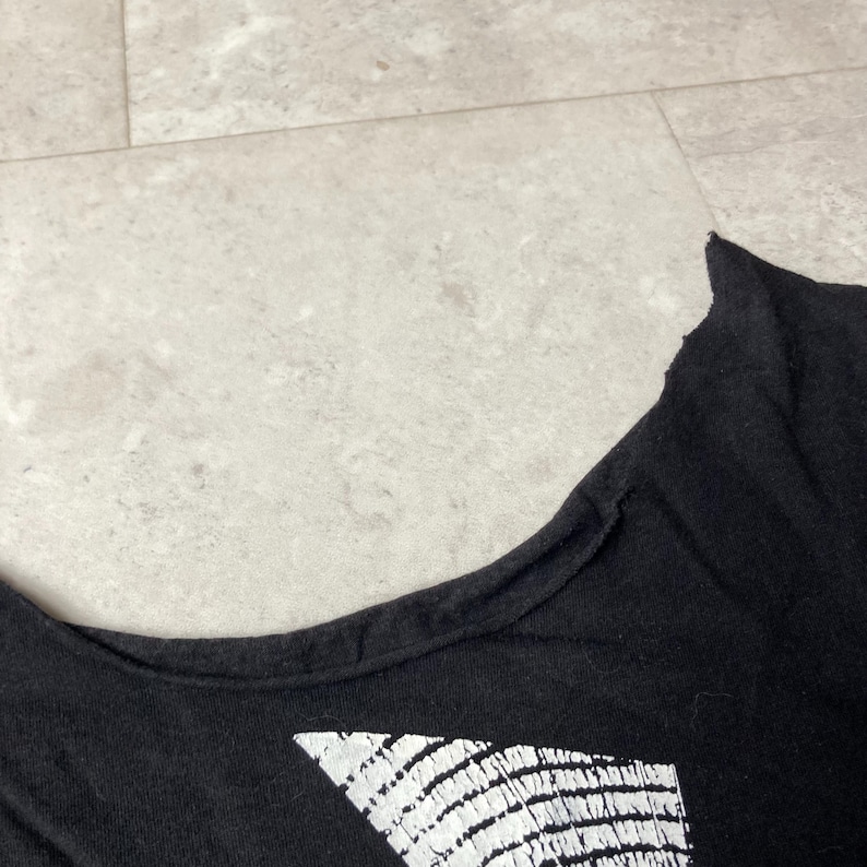 May include: Black t-shirt with a white graphic design. The shirt has a scoop neck and a distressed, raw edge along the neckline. The graphic appears to be a geometric pattern.