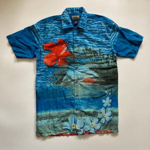 May include: Blue short-sleeve button-down shirt with a tropical print. The shirt features a large red hibiscus flower, a sunset scene, and white and blue floral accents. The shirt has a collared neckline and a button-up front.