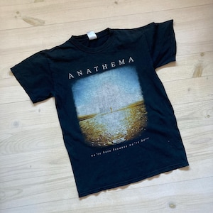 May include: Black t-shirt featuring the band name "ANATHEMA" in white block letters above a rectangular image of a person standing in water. The text "we're here because we're here" is below the image.