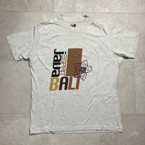Può includere: T-shirt grigio chiaro con un design grafico. Il design include le parole "JAVA" e "BALI" in vari colori, il marchio "ESPRIT" e un'illustrazione floreale. La maglietta ha maniche corte e girocollo.