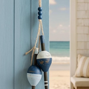 Op de afbeelding: Decoratieve hangende boei-ornamenten met een nautisch thema. De boeien zijn geschilderd in blauw- en wittinten, met een verweerde afwerking. Ze zijn opgehangen aan een natuurlijk touw en blauwe houten kralen. De achtergrond toont een strandscène.