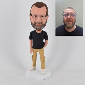 May include: A custom bobblehead figurine of a man with a beard and glasses, wearing a black t-shirt and tan cargo pants. The figurine is on a white base, with a photo of the man in the background.