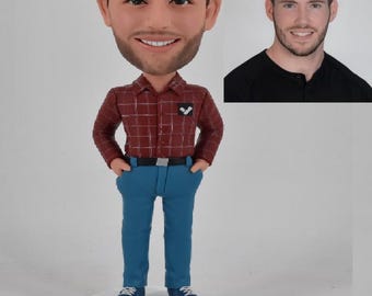 Puede incluir: Figura de bobblehead personalizada de un hombre con una camisa a cuadros granate, pantalones azules y zapatos azules. La figura tiene una cara sonriente y está sobre una base blanca. Una foto del hombre está en el fondo.
