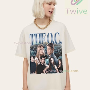 Puede incluir: Camiseta color crema con un gráfico de la serie de televisión "The O.C." con una foto del elenco y el título del programa en azul. La camiseta la lleva una persona con cabello rubio y un collar de cadena dorada.