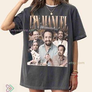May include: Dark gray t-shirt featuring a collage of images of Lin-Manuel Miranda with the text "Lin-Manuel Miranda" in gold lettering. The shirt has a vintage, slightly faded appearance.