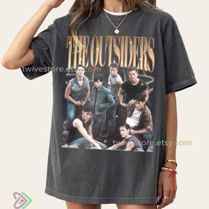 May include: Dark gray t-shirt featuring a graphic of the movie "The Outsiders" with a group of actors. The text "THE OUTSIDERS" is in gold lettering. The Etsy shop name "twivestore.etsy.com" is printed on the shirt.