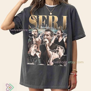 May include: Dark gray t-shirt featuring a collage of a singer with a microphone, with the name "SERJ" in gold lettering. The shirt has the text "Tankian" and the Etsy shop name "twivestore.etsy.com".