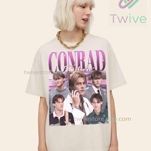 May include: Cream-colored t-shirt featuring a graphic of Conrad Fisher with a vintage aesthetic. The design includes the name "CONRAD Fisher" in a purple font, along with multiple images of the person. The shirt is worn by a person with blonde hair and a gold chain.