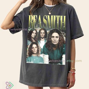 May include: Dark gray t-shirt featuring the name "BEA SMITH" in gold lettering above a collage of four images of a woman. The images are in a rectangular frame. The shirt has short sleeves and a relaxed fit.