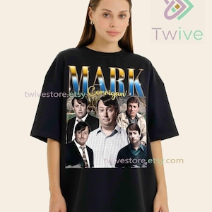 Peut inclure: T-shirt noir avec un graphisme doré et bleu du texte "MARK Corrigan" et un collage d'images du personnage. Le t-shirt est de style col rond classique.