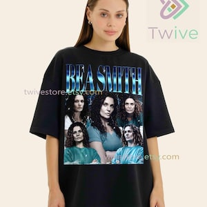 May include: Black t-shirt featuring the name "BEA SMITH" in a blue gradient font. The shirt displays multiple images of a woman with curly hair. The Twive logo is visible in the upper right corner.