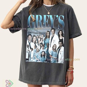 Könnte beinhalten: Dunkelgraues T-Shirt mit dem Text "GREY'S Anatomy" in einem blauen Farbverlauf über einem Gruppenfoto der Besetzung der Serie. Das Shirt hat einen Vintage-Look mit der Etsy-Shop-URL auf der Seite.
