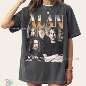May include: Dark gray t-shirt featuring the name "ALAN" in gold and orange lettering, with a photo of Alan Rickman. The shirt has a vintage style and includes the name "Rickman" below the photo.
