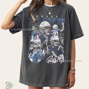 May include: Dark gray t-shirt featuring a collage of images of Drake Maye in a football uniform. The design includes the name "Drake Maye" in blue and white lettering. The shirt has a vintage, worn look.