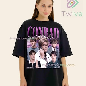 May include: Black t-shirt featuring a collage of images of a person, with the name "CONRAD Fisher" in a purple and white design. The shirt has a crew neck and short sleeves. The Twive logo is in the upper right corner.