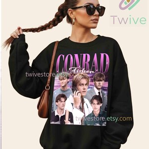 May include: Black sweatshirt featuring a graphic of the name "CONRAD Fisher" in a pink and purple gradient font, with multiple images of a person. The sweatshirt has long sleeves and a crew neck. The Etsy shop name "twivestore.etsy.com" is visible.
