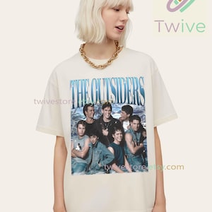 May include: Cream-colored t-shirt featuring a group photo and the text "THE OUTSIDERS" in blue. The shirt has short sleeves and a crew neck. The image is a vintage-style design.