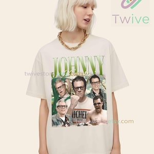 May include: Cream-colored t-shirt featuring a graphic of Johnny Knoxville and other men. The design includes the name "Johnny Knoxville" in green, with a vintage aesthetic. The shirt is worn by a person with short blonde hair and a gold chain.