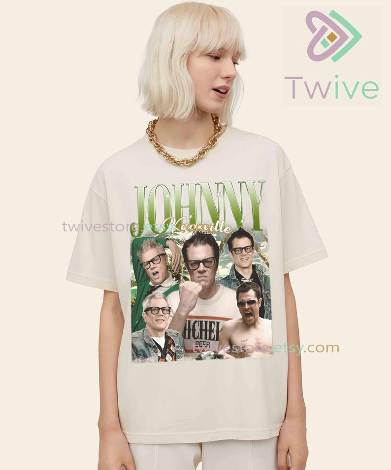 May include: Cream-colored t-shirt featuring a graphic of Johnny Knoxville and other men. The design includes the name "Johnny Knoxville" in green, with a vintage aesthetic. The shirt is worn by a person with short blonde hair and a gold chain.
