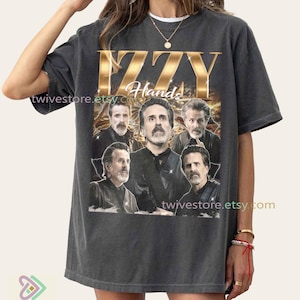 May include: Dark gray t-shirt featuring a gold-colored "IZZY Hands" graphic with a collage of images of a man. The Etsy shop URL "twivestore.etsy.com" is printed on the shirt. The shirt has a vintage, worn look.