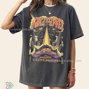 May include: Dark gray t-shirt featuring a colorful graphic of a lizard-like creature breathing fire over a city, with the text "KING GIZZARD AND THE LIZARD WIZARD". The shirt has a vintage, worn look.