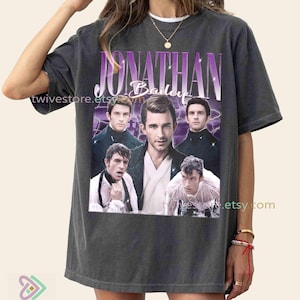 May include: Dark gray t-shirt featuring a graphic of Jonathan Bailey with the name in a purple font. The design includes multiple images of the actor. The Etsy shop name is visible on the shirt.