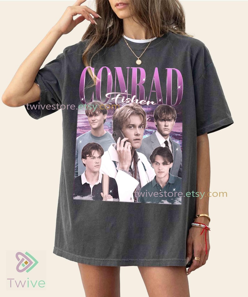 May include: Dark gray t-shirt featuring a graphic of a person with the text "CONRAD Fisher" in a pink and white design. The shirt has a vintage style and includes multiple images of the person.