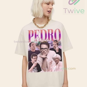 May include: Cream-colored t-shirt featuring a graphic of Pedro Pascal with the text "PEDRO Pascal". The design includes multiple images of the actor. The shirt is worn by a person with blonde hair and a gold chain necklace.