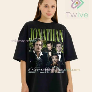 Puede incluir: Camiseta negra con un diseño de Jonathan Groff y el nombre "Jonathan" en verde. El diseño incluye varias imágenes del actor. La camiseta tiene un corte holgado.