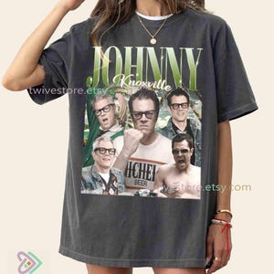 May include: Dark gray t-shirt featuring a collage of images of Johnny Knoxville and the text "JOHNNY Knoxville" in green and white. The shirt has a vintage look and feel.