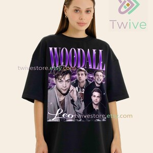 May include: Black t-shirt featuring a graphic of the name "WOODALL" in purple, with a collage of images of a person. The name "Leo" is written in cursive below the images. The shirt has the website address "twivestore.etsy.com" printed on it.