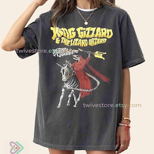 May include: Charcoal grey t-shirt with the band name "King Gizzard & The Lizard Wizard" in yellow. The graphic depicts a skeleton in a red cloak on a skeletal horse, holding a guitar. The shirt also has the text "October 12 2018 Tijuana".