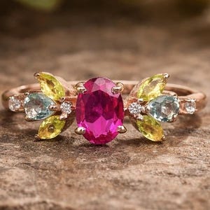 Puede incluir: Un anillo de oro rosa con una gema central magenta de corte ovalado. El anillo está adornado con gemas más pequeñas en amarillo, azul y blanco, formando un diseño floral. Se muestra sobre una superficie marrón texturizada.