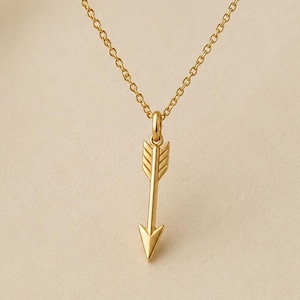 May include: A gold-toned necklace featuring a delicate chain and a vertical arrow-shaped pendant. The pendant displays a textured fletching design at the top and a pointed tip at the bottom, suspended from a small circular jump ring.