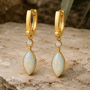 May include: A pair of gold hoop earrings featuring a small round clear stone and a marquise-shaped white opal cabochon drop. The earrings have a polished gold finish and a hinged closure mechanism, showcasing a clean and symmetrical design.