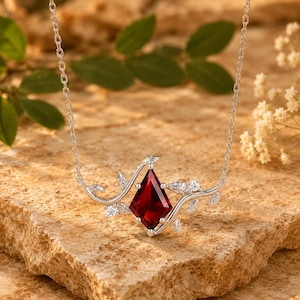 May include: A silver-toned necklace featuring a kite-shaped deep red gemstone centerpiece. The setting is designed with delicate metal vine and leaf motifs extending from the stone, accented by small clear round stones along the branches.