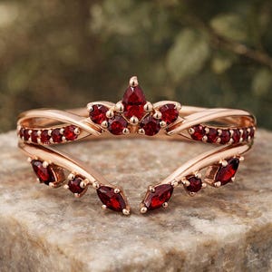 May include: A rose gold ring featuring a curved, open-ended band design adorned with multiple deep red, faceted gemstones. The stones are arranged in marquise and round shapes along the top and sides of the metal band, creating a symmetrical look.