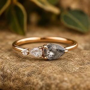May include: A rose gold ring featuring a cluster of three stones: a small round clear stone, a pear-shaped clear stone, and a larger pear-shaped grey rutilated quartz stone with visible dark needle-like inclusions set in a polished metal band.