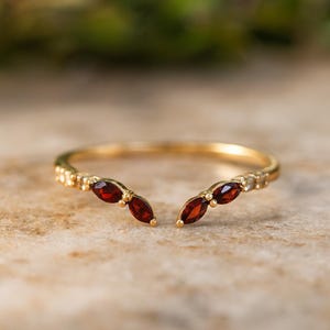 Puede incluir: Un anillo abierto de oro con dos piedras granates de color rojo intenso en talla marquesa en cada extremo, acentuado por pequeñas piedras claras a lo largo de la banda. El anillo tiene una estructura de metal fina y delicada con acabado pulido.