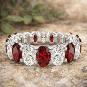 May include: A silver eternity band featuring alternating oval-cut red gemstones and clear white gemstones set in a continuous row. The metal band has a polished finish, and each stone is secured by four prongs, creating a repeating pattern around the ring.