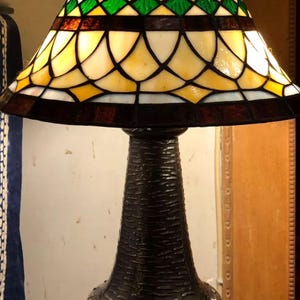 May include: A stained glass table lamp with a bronze base. The lampshade features a geometric pattern of yellow, green, and brown glass. The base has a textured, dark brown finish. The lamp is designed for indoor use and provides ambient lighting.