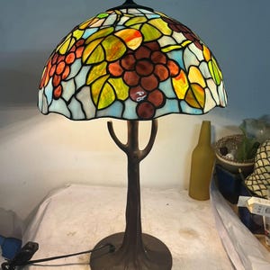 Puede incluir: Una lámpara de mesa de vidrieras con una pantalla abovedada que presenta un diseño de racimo de uvas en tonos de rojo, naranja, amarillo y verde. La lámpara tiene una base y un tallo de color bronce, que se asemejan a un árbol. La lámpara mide aproximadamente 51 cm de altura.