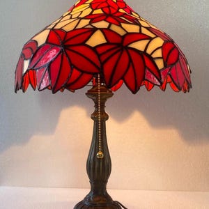 Può includere: Lampada da tavolo con paralume in vetro colorato. Il paralume presenta un motivo floreale rosso e giallo. La base è color bronzo e la lampada ha una catena. Adatta per l'illuminazione interna.