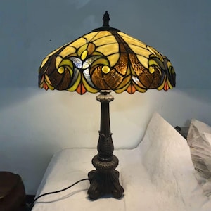 May include: A stained glass table lamp with a bronze base. The lampshade is a mosaic of yellow, brown, and orange glass pieces. The lamp is turned on, casting a warm glow. The lamp is on a white surface.
