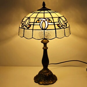 Puede incluir: Una lámpara de mesa decorativa con una pantalla de vidriera. La pantalla es de color crema con un patrón geométrico y un borde festoneado. La base es de color bronce con un diseño detallado. La lámpara tiene un cable de alimentación negro.