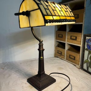 May include: A desk lamp with a stained glass shade, featuring a bronze base and a curved design. The shade is made of yellow and amber glass pieces, with a black border. A pull chain is visible.