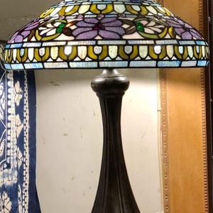May include: A stained glass table lamp with a bronze base. The lamp shade features a floral design in shades of purple, blue, green, and yellow. The lamp is turned on, illuminating the colourful glass.