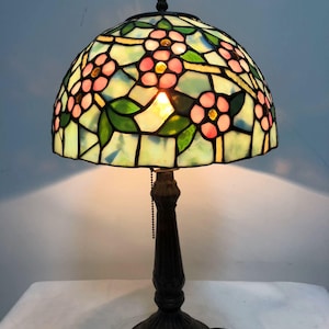 May include: A decorative table lamp with a stained glass lampshade. The lampshade features pink flower motifs, green leaves, and a light blue background. The base is dark brown and ornate, with a pull chain.