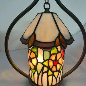 May include: A decorative stained glass lamp with a floral pattern. The lamp has a white top and a body made of colourful glass pieces in red, orange, green, and yellow. It hangs from a dark metal frame.